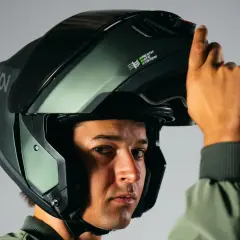 Un hombre se ajusta un casco de moto verde oscuro, similar a un casco guardia civil, con el sistema "Breath Safe" visible.