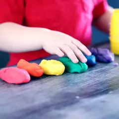Dziecko w czerwonej koszulce bawi się plasteliną w tęczowych kolorach. To idealny obrazek do materiałów o kursie na opiekuna w żłobku.