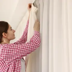 Kobieta w kratę wiesza białe firanki na suficie, pokazując, jak powiesić firanki bez karnisza.