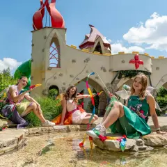 Trzy dziewczynki w strojach wróżek siedzą nad wodą w bajkowym parku rozrywki dla dzieci.
