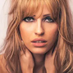 Kobieta z blond włosami i cieniowaniem grzywki, z dłońmi na szyi.