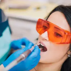 Pacjentka w okularach ochronnych poddaje się zabiegowi stomatologicznemu. Dentysta w rękawiczkach używa wiertła. Nowoczesne znieczulenie komputerowe zapewnia komfort.