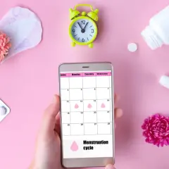 Telefon z kalendarzem cyklu menstruacyjnego, obok podpaska, tampon, leki i kwiaty. Warto pamiętać o mięśniakach macicy.