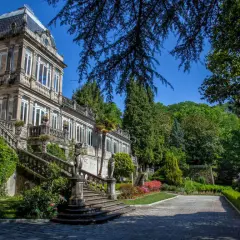 El Pazo de Lourizán, una mansión señorial con escalinatas cubiertas de hiedra y un jardín exuberante, se alza majestuoso bajo un cielo azul.