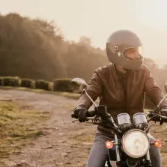 Mężczyzna w kasku i skórzanej kurtce jedzie na motocyklu po polnej drodze. Słońce zachodzi, tworząc ciepłą, złotą poświatę.