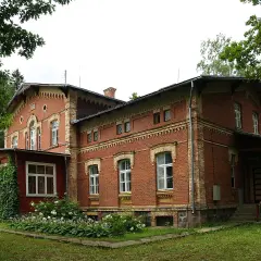 Stary, ceglany maison otoczony zielenią, z werandą i kwitnącymi krzewami.