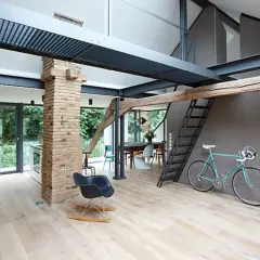 Moderner offener Dachstuhl mit sichtbaren Stahlträgern, Ziegelpfeiler, Holz und einer Leiter. Ein Fahrrad steht bereit.