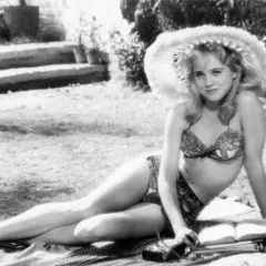 Młoda kobieta w bikini i kapeluszu leży na trawie, czytając książkę. Scena jak z letnich filmów erotycznych.