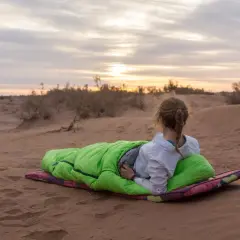 Fin du **trek dans le désert**. Une jeune femme en sac de couchage vert admire le coucher de soleil sur les dunes.