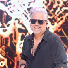 Artur Gadowski, wokalista zespołu IRA, w czarnej koszuli i okularach przeciwsłonecznych, uśmiecha się na tle kolorowej scenografii.