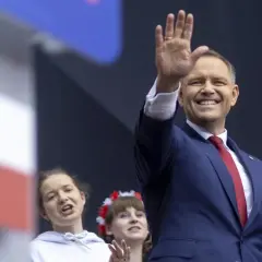 Uśmiechnięty polityk w garniturze pozdrawia tłum. W tle dwie młode kobiety, jedna z wiankiem na głowie. To może być część kampanii wyborczej, pozew w trybie wyborczym.
