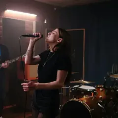Wokalistka śpiewa do mikrofonu, basista gra na gitarze, a perkusista wybija rytm w muzyce.