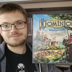 Uśmiechnięty mężczyzna prezentuje pudełko gry "Dominion II Edycja". To dopiero początek przygody z nowymi dodatkami!
