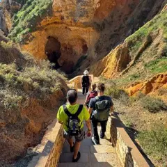 Hikers descend a stone staircase on a scenic trail in Portugal, leading towards a large cave entrance nestled within rugged, sun-drenched cliffs.