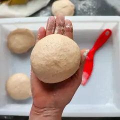 Hand hält klebrigen Pizzateig. Mehr Teiglinge in einer weißen Schale, daneben ein roter Teigschaber.