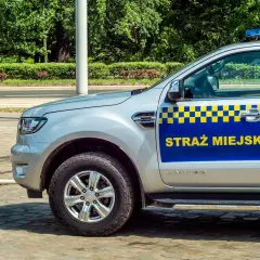 Srebrny samochód Straży Miejskiej z niebiesko-żółtym pasem i napisem "STRAŻ MIEJSKA".