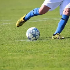 Piłkarz w niebieskich getrach i żółtych butach wykonuje rzut rożny. Piłka leży na trawie, gotowa do kopnięcia.