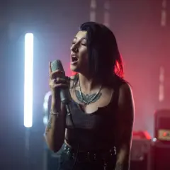 Młoda kobieta z tatuażami, w rockowym stylu, z pasją wykonuje śpiew operowy do mikrofonu. W tle neonowe światła i wzmacniacz Marshall.