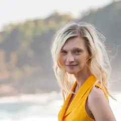 Agata Buzek w żółtej sukience, z falującymi blond włosami, uśmiecha się na tle morza i wzgórz.