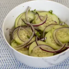 Deliciosa ensalada de pepino con cebolla morada y hierbas frescas, servida en un tazón blanco sobre un mantel azul y blanco.