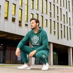 Mężczyzna w dresie butelkowa zieleń z nadrukiem "College" kuca przed nowoczesnym budynkiem z żółtymi oknami.