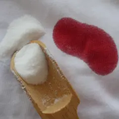Une cuillère en bois contient une poudre blanche à côté d'une tache rouge sur un tissu blanc. Une tache de sang qui ne part pas au lavage, c'est le cauchemar de tout parent.