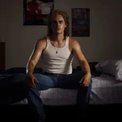 Billy z Stranger Things siedzi na łóżku w białej podkoszulce i dżinsach, w pokoju z plakatami na ścianie.