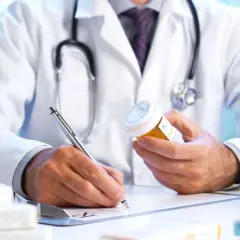 Medico con camice bianco e stetoscopio, scrive su ricetta medica, tenendo in mano un flacone di pillole. Un piano terapeutico medico di base in corso.