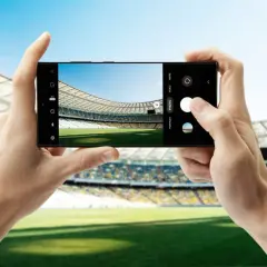 Dwie dłonie trzymają smartfon Samsung S23, robiąc zdjęcie pustego stadionu piłkarskiego.