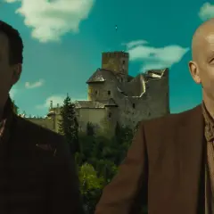 Dwóch mężczyzn, jeden z nich łysy, stoi przed zamkiem. Słońce oślepia ich od świateł.