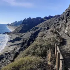 Sendero costero con vistas al mar Mediterráneo y montañas. Un camino de tierra con barandilla de madera serpentea por la ladera, ideal para un paseo marítimo en Mojácar Playa.
