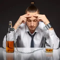 Mężczyzna w kajdanach, przykuty do butelki i szklanki z alkoholem, symbolizuje walkę z nałogiem. Suchy alkoholik, który mimo abstynencji, wciąż czuje ciężar uzależnienia.