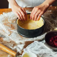Dłonie formują ciasto w tortownicy na drewnianym blacie z mąką.