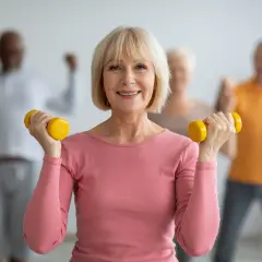 Une femme souriante fait de la gymnastique senior avec des haltères jaunes. D'autres personnes âgées s'entraînent en arrière-plan.