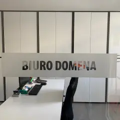Szklana witryna z napisem "BIURO DOMENA" na tle nowoczesnego biura z białymi szafami.