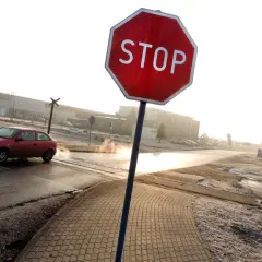 Czerwony samochód zbliża się do skrzyżowania, gdzie widoczny jest znak stop. W tle budynki przemysłowe i kolejowy przejazd.