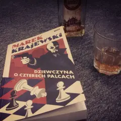 Powieść Marka Krajewskiego "Dziewczyna o czterech palcach" leży obok szklanki z bursztynowym trunkiem.