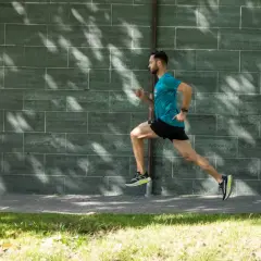 Mężczyzna w turkusowej koszulce i czarnych spodenkach wykonuje bieg ciągły wzdłuż zielonej, ceglanej ściany.