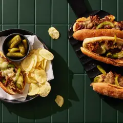 Två philly cheese steak-mackor med smält ost, paprika och lök. Serveras med cornichoner och potatischips.