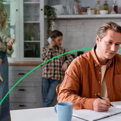 Mężczyzna w pomarańczowej koszuli pracuje przy laptopie, podczas gdy rodzina jest w tle.