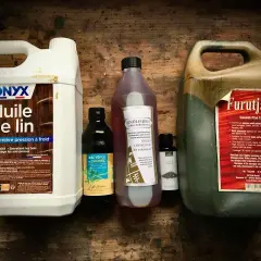 Plusieurs bouteilles d'huile pour le bois, dont de l'huile de lin et du goudron de pin, sont alignées sur une surface en bois.