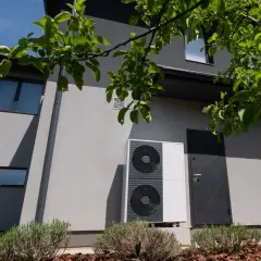 Unità esterna di una pompa di calore residenziale, moderna e discreta, installata accanto a una porta d'ingresso su un edificio grigio.