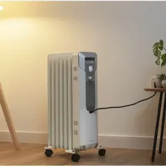 Estufa eléctrica de aceite con ruedas, conectada a la corriente, junto a una planta y una lámpara. Controla el consumo.