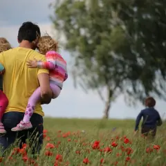 Un padre porta in braccio le sue due figlie in un campo di papaveri, un'immagine che cattura la psicologia del rapporto padre e figlia.