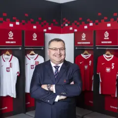 Uśmiechnięty mężczyzna w garniturze stoi przed szatnią reprezentacji Polski. Widać tam koszulki z nazwiskami znanych piłkarzy, tworząc zgrupowanie reprezentacji Polski.