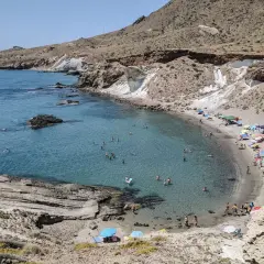 Bahía de aguas cristalinas con gente bañándose y disfrutando de las **cabo de gata playas**. Sombrillas de colores y arena dorada bajo un cielo azul.