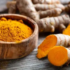 Raíces de cúrcuma y polvo dorado en cuenco de madera, evocando las propiedades de la cúrcuma en cápsulas.