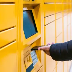 Ręka zbliża smartfon do żółtego paczkomatu InPost, aby sprawdzić, ile paczka czeka w paczkomacie.