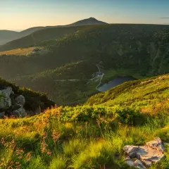 Widok na malowniczy szczyt w Karkonoszach o zachodzie słońca. W dole jezioro i schronisko, otoczone zielonymi zboczami.