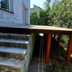 Nowy taras na słupach stalowych, z drewnianą podłogą, sąsiaduje z betonowymi schodami i zielonym ogrodem.